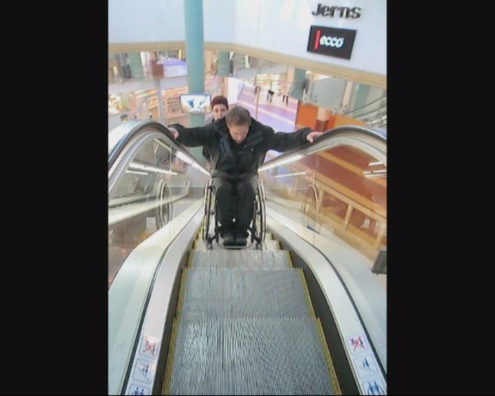 User rides down an escalator with help from an assistant.
